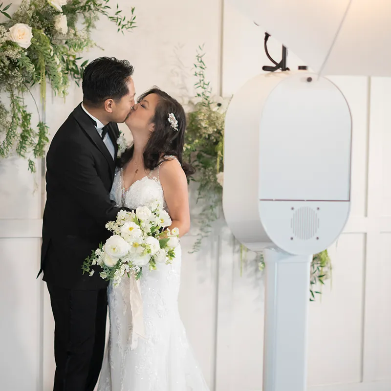 Bride and groom kissing inside an Olive Branch photo booth holding props.